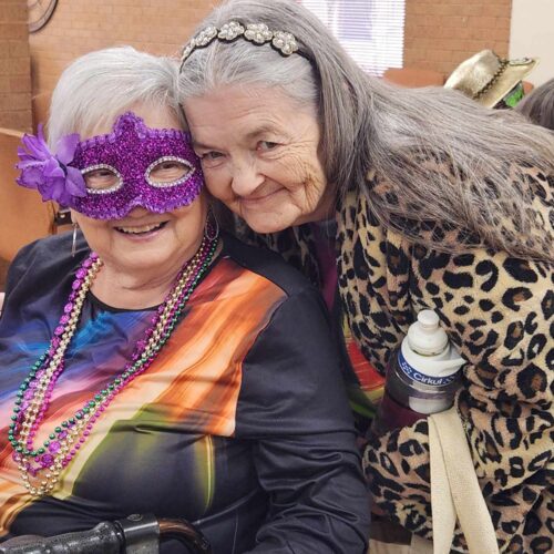 Woman leans down to hug woman in wheelchair with mardi gras mask on.