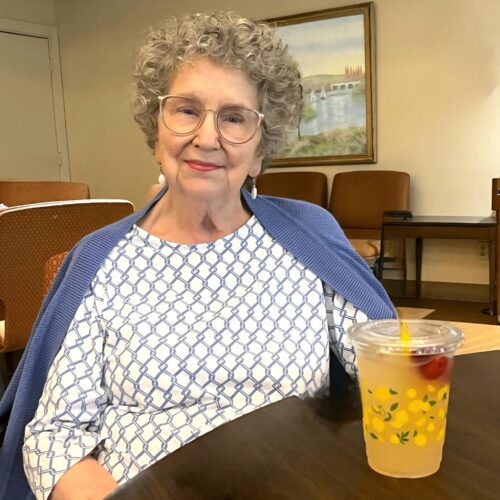 An elderly woman with curly grey hair and large glasses, wearing a blue and white patterned shirt, smiling while sitting at a wooden table with a drink.