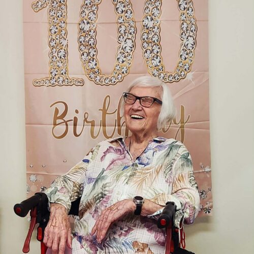 An elderly woman smiling with a decorative "100th Birthday" sign while celebrating at Good Shepherd Community.
