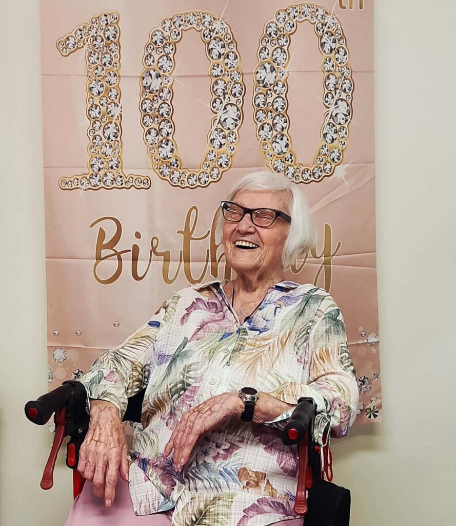 An elderly woman smiling with a decorative "100th Birthday" sign while celebrating at Good Shepherd Community.